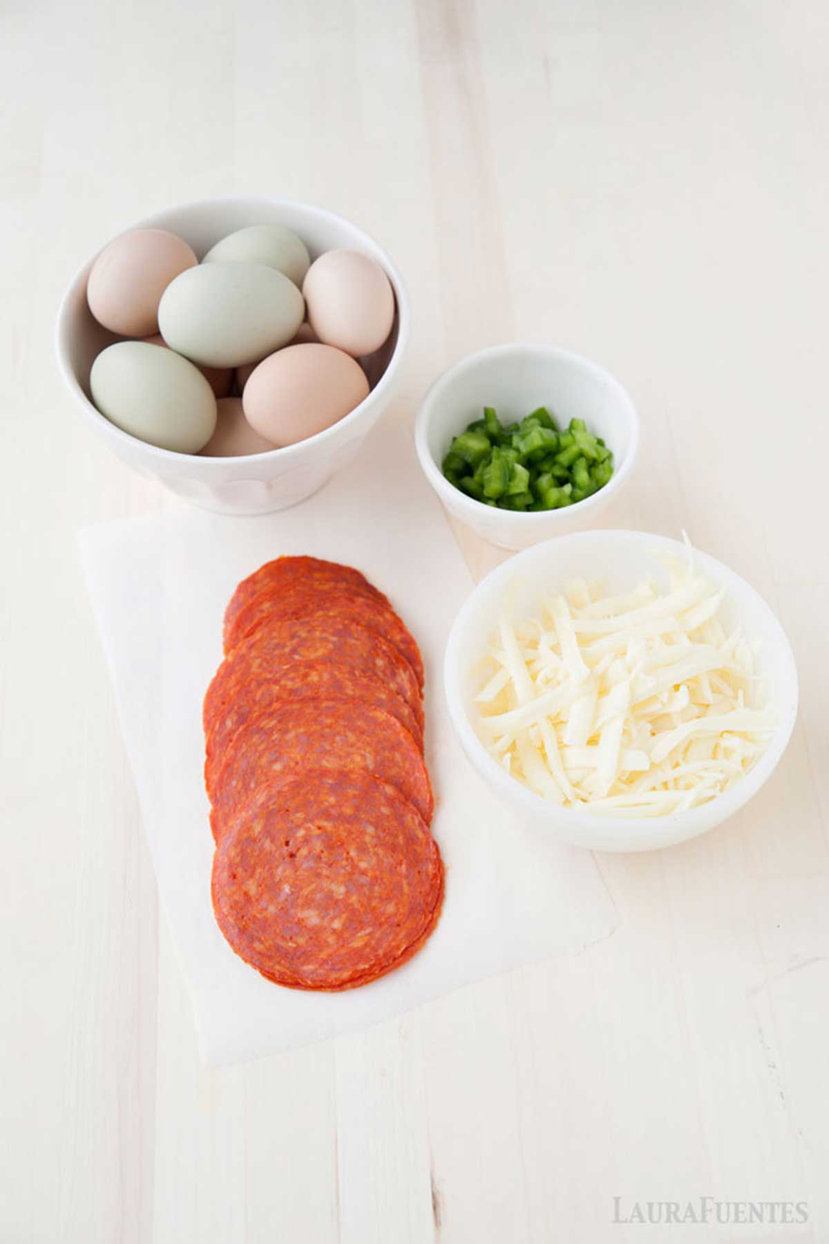 un bol con huevos, otros con pimiento verde y otro con queso rallado. Sobre la mesa, rodajas de salami.