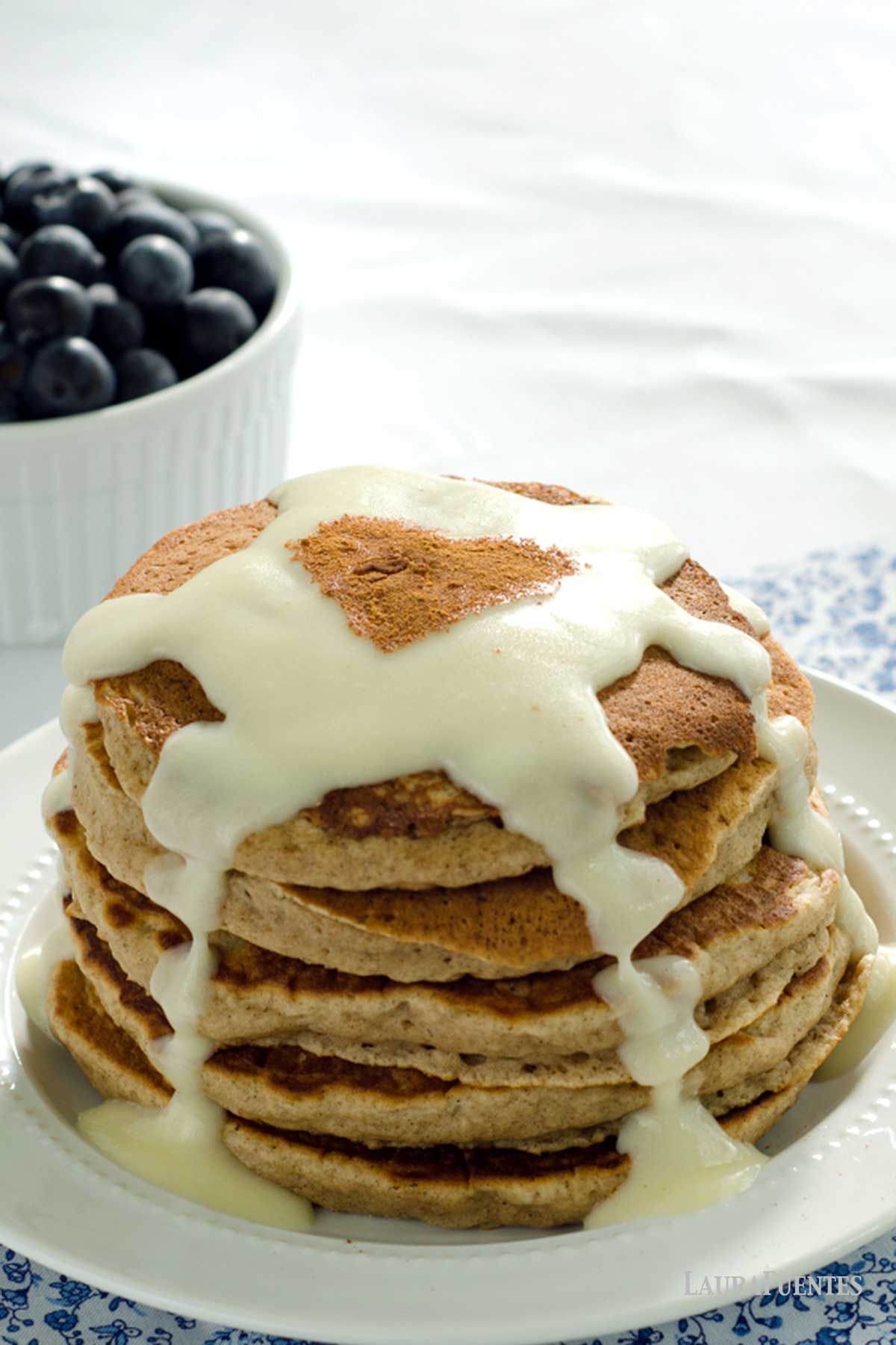 una pila de pancakes de canela con glaseaso sobre un plato, con un bol de arándanos detrás.