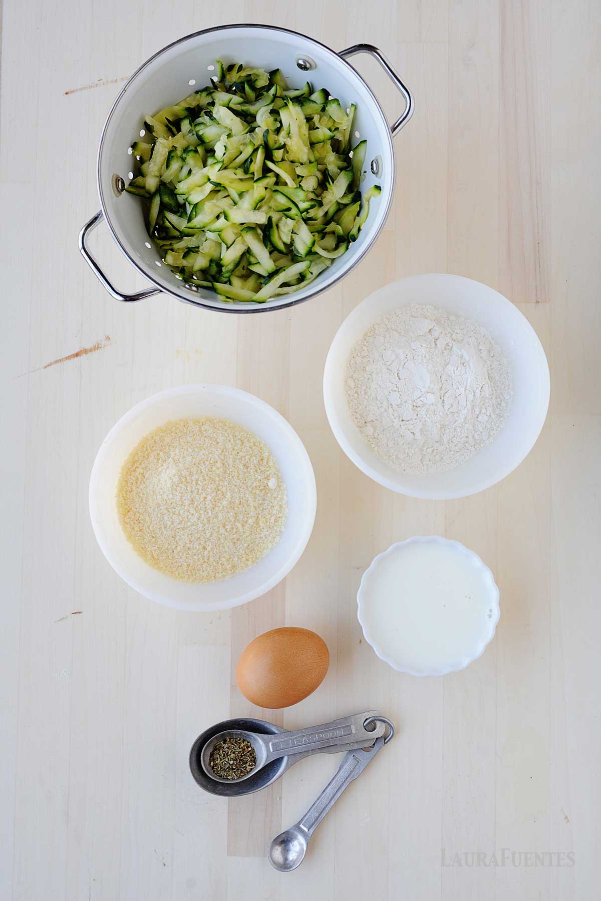 calabacín rallado en una olla, bols con queso, harina y leche, un huevo y unas cucharas medidoras