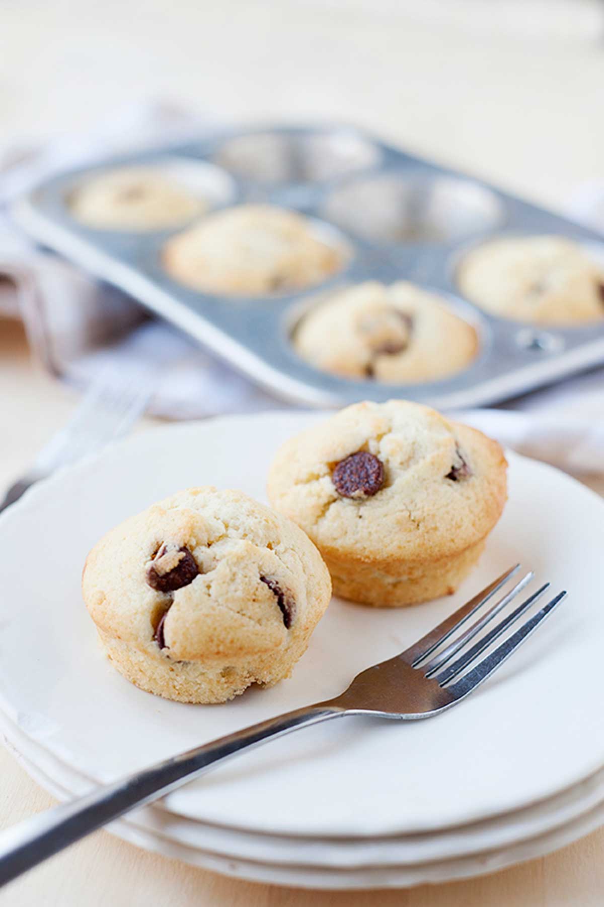 dos muffins con chispas de chocolate en un plato con un tenedor. Detrás, una placa para muffins