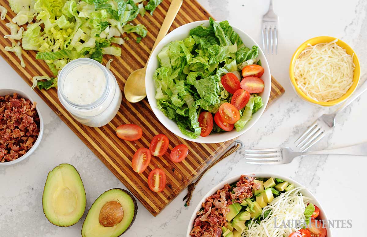 un bol con lechuga y tomate, otro con queso rallado, otro con ensalada y otro con bacon cocido. También  un aguacate, tomates, un frasco de salsa ranch y varios cubiertos.
