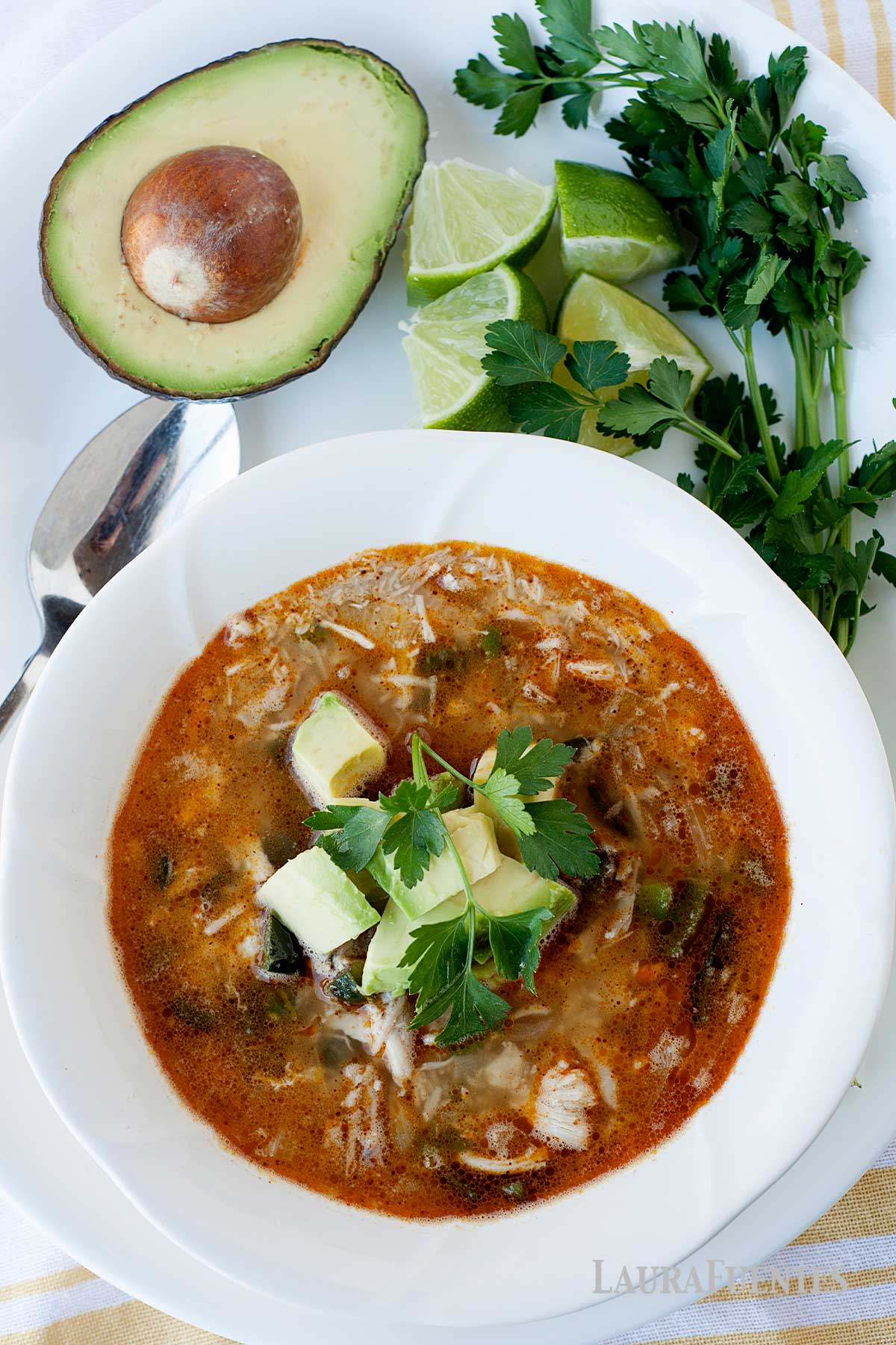 un plato de sopa de pollo desmenuzado, junto a un aguacate y rodajas de lima