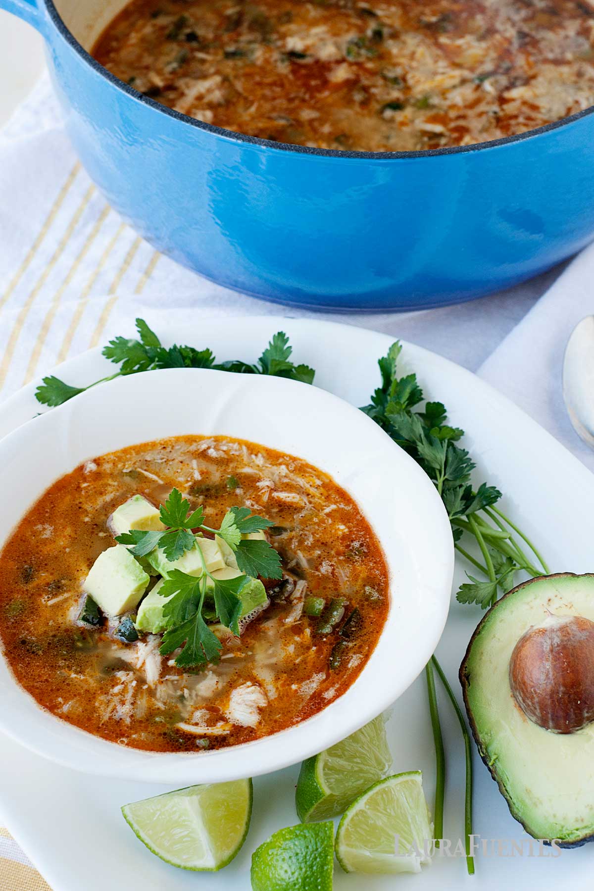 un plato y una olla con sopa de pollo desmenuzado, junto a un aguacate y rodajas de lima