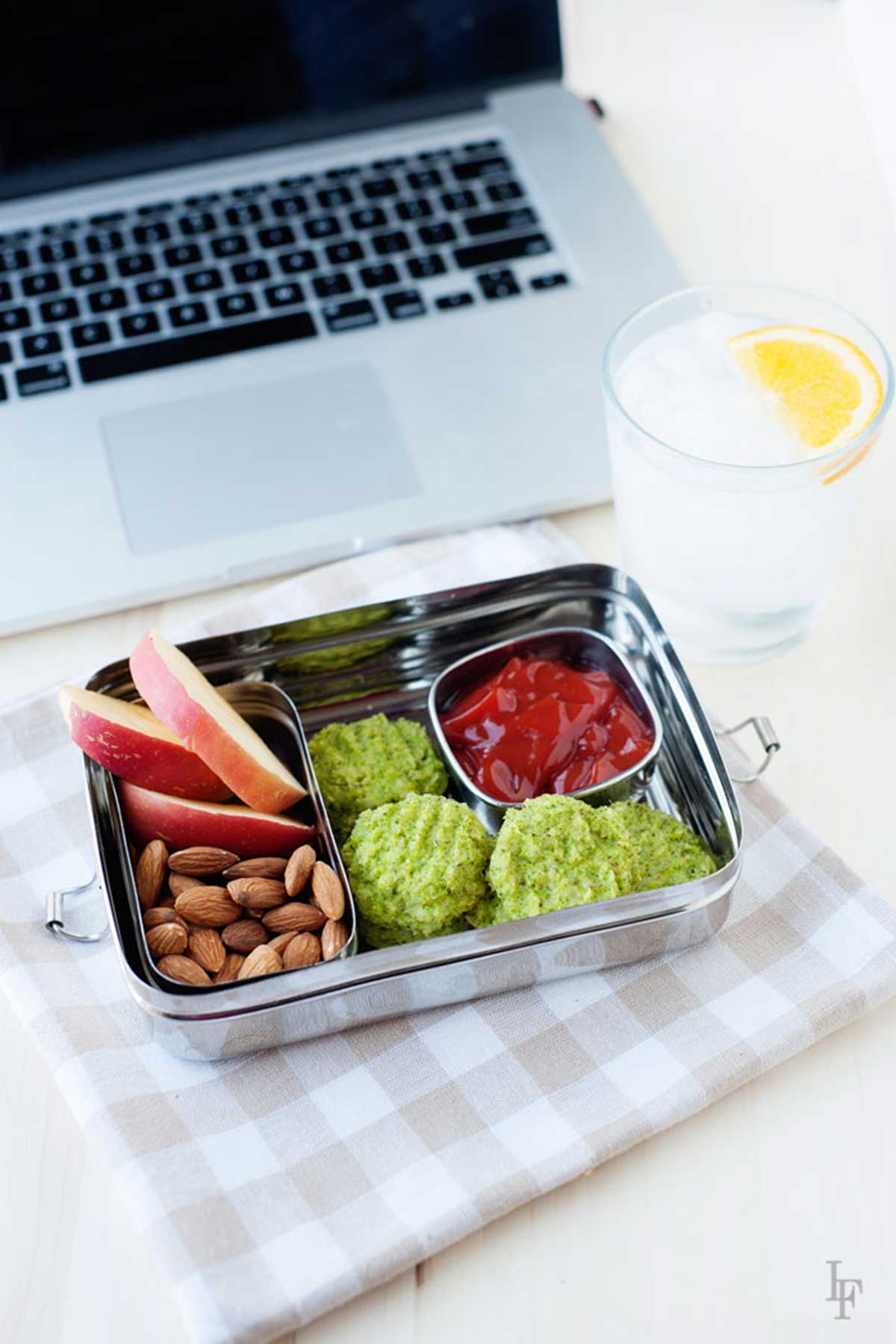 ordenador, vaso de agua, nuggets verdes de brocoli y queso, almendras y manzana en un contenedor metal.