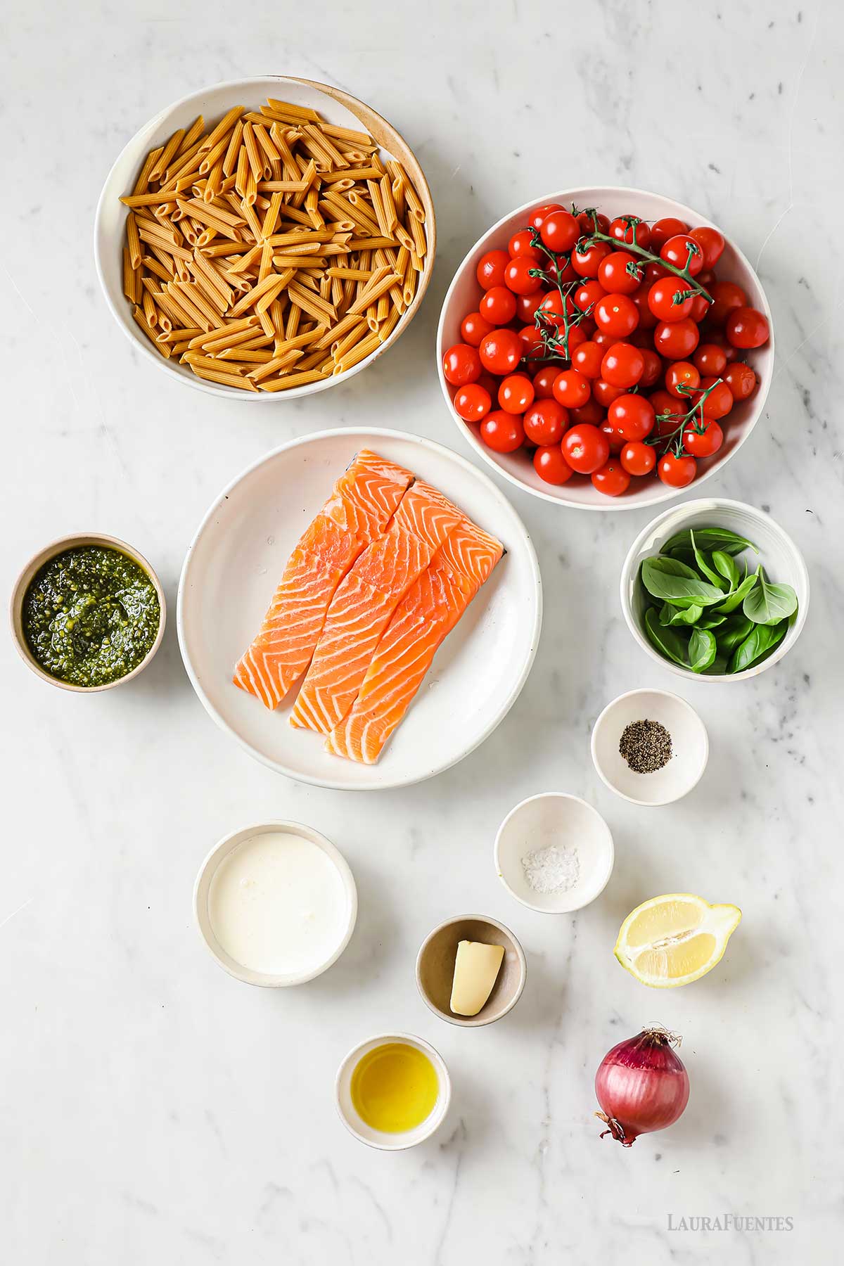 Bols de distintos tamaños con los ingredientes de la receta: pasta, tomates pequeños, salmón, pesto, albahaca, aceite, sal, pimienta, nata, mantequilla, junto a medio limón y una cebolla.