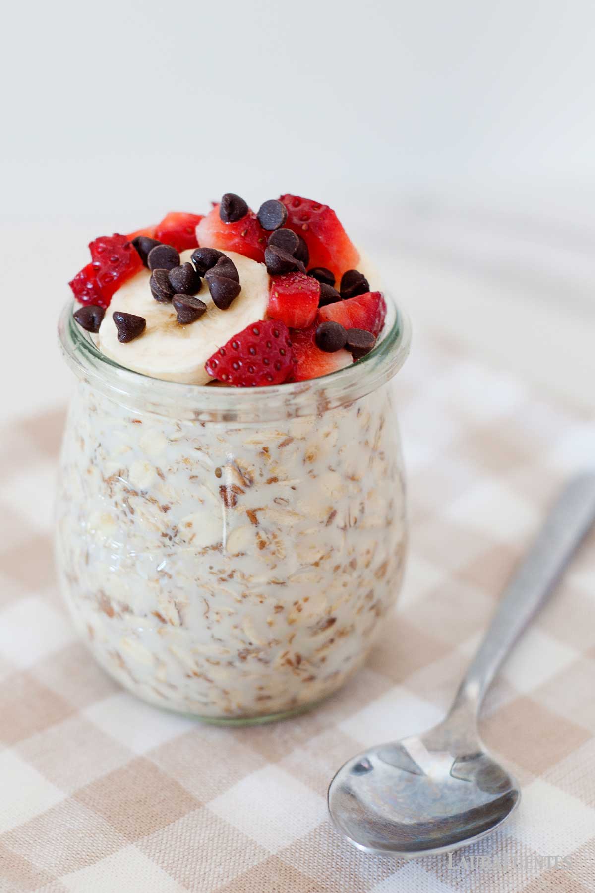 un frasco de avena con leche, plátano, fresas y chispas de chocolate, con una cuchara al lado.