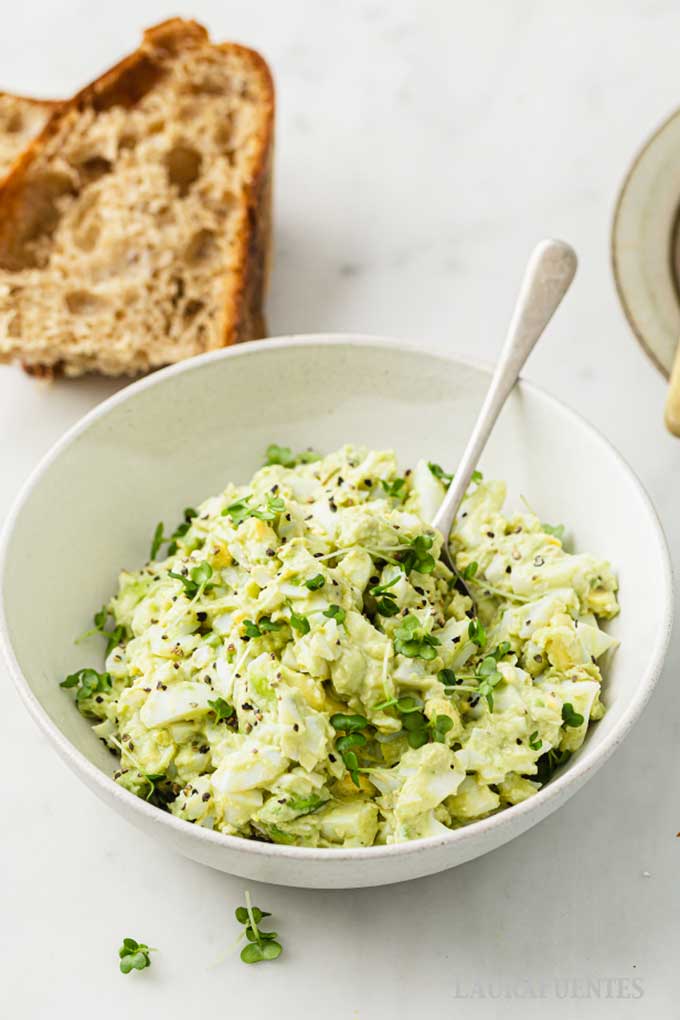 un bol de ensalada de aguacate con huevo duro y un trozo de pan detrás