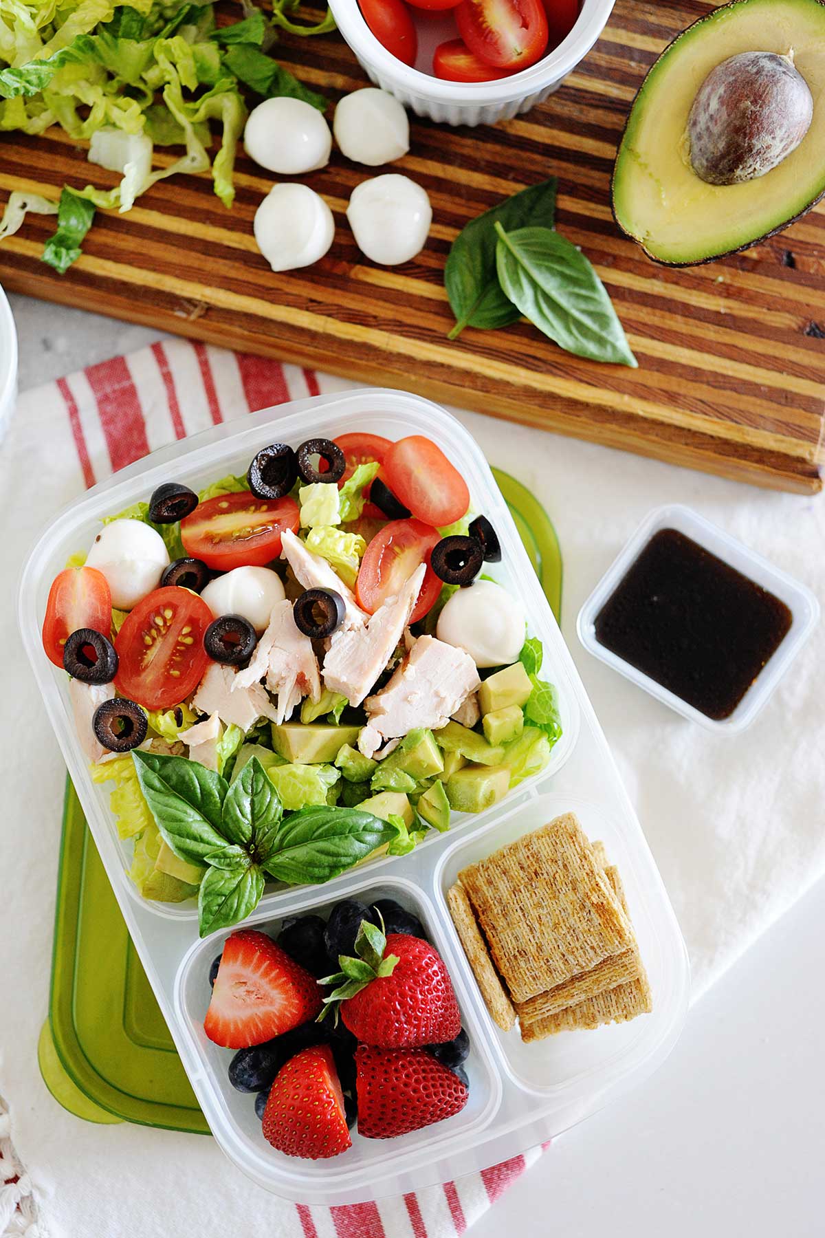 un hermético para llevar con ensalada de tomate, pollo, aceitunas, lechuga, crackers, frutas y un pequeño recipiente con vinagreta