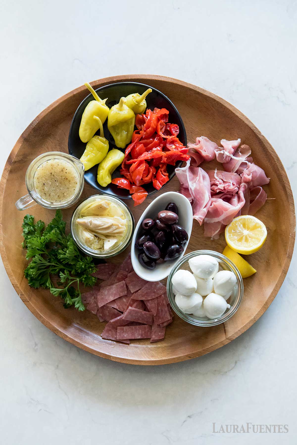 una tabla con salami, limón, jamón serrano, aceitunas, corazones de alcachofa, vinagreta de mostaza, mozzarella, perejil, pimientos rojos asados y peperonccini