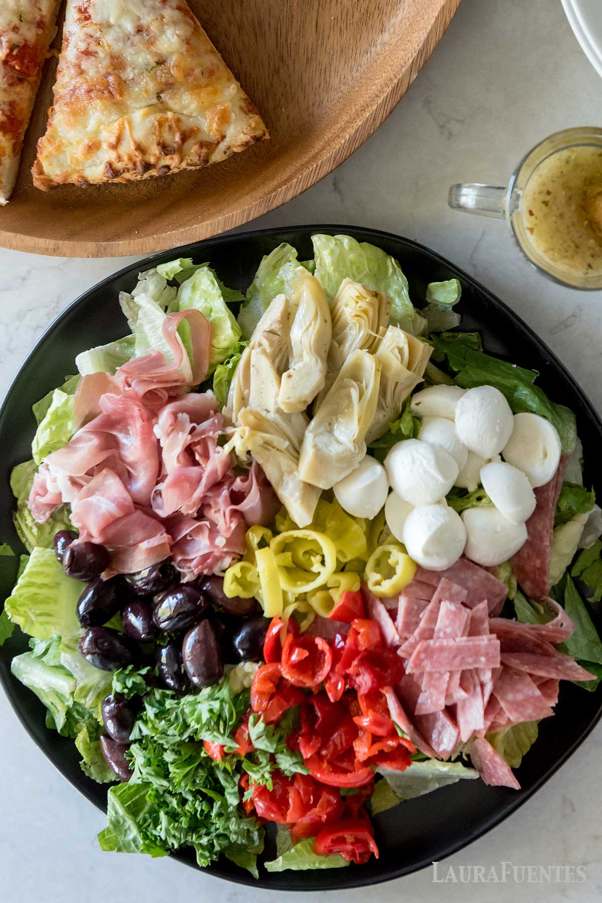 una fuente con ensalada antipasto junto a una tabla con pizza y una taza.
