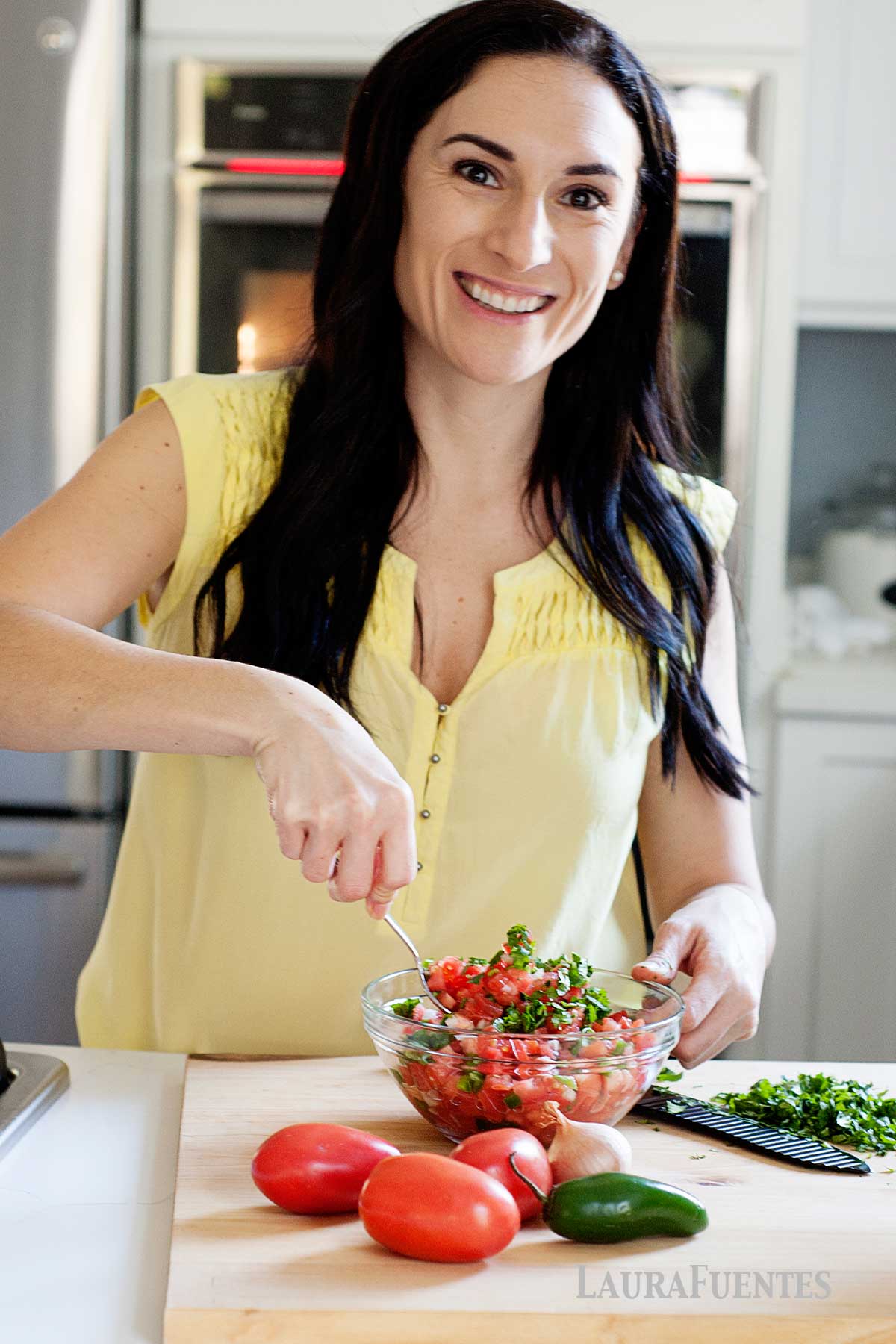 laura fuentes revolviendo un bol con pico de gallo sobre una mesa donde también hay jalapeño, cebolla, tomates y cilantro.