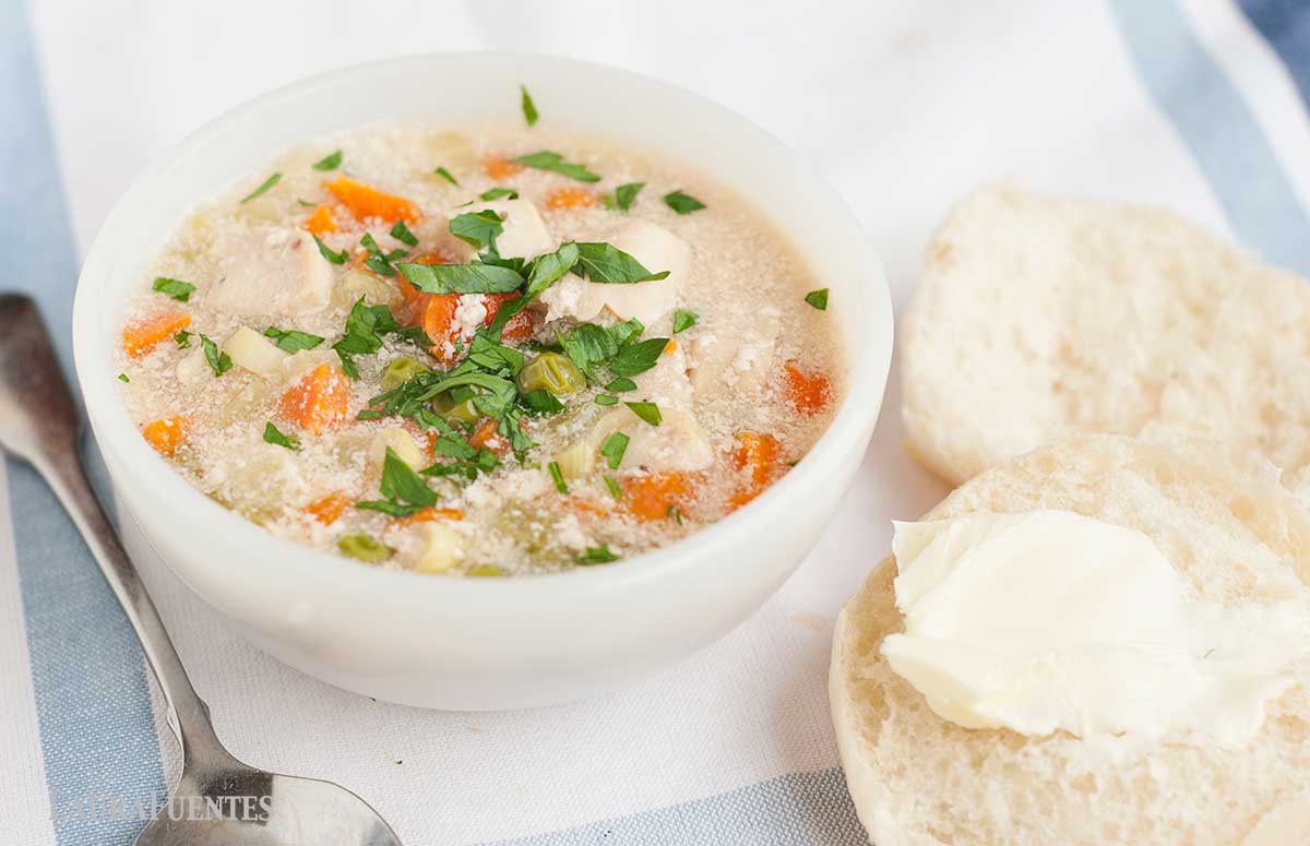 un tazón de sopa de verduras y pollo con una cuchara dentro y dos panecillos al lado