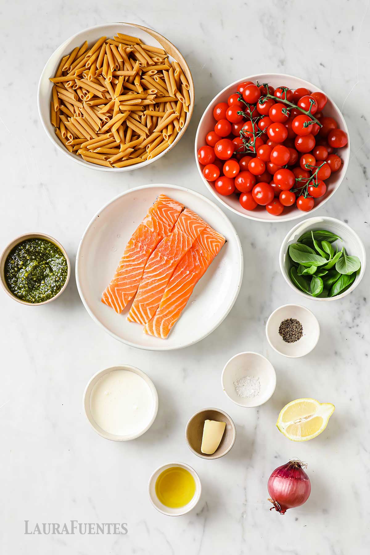 varios recipientes con pasta cruda, tomates cherry, salmón, albahaca, pesto, aceite y condimentos. Al lado, una cebolla y medio limón.