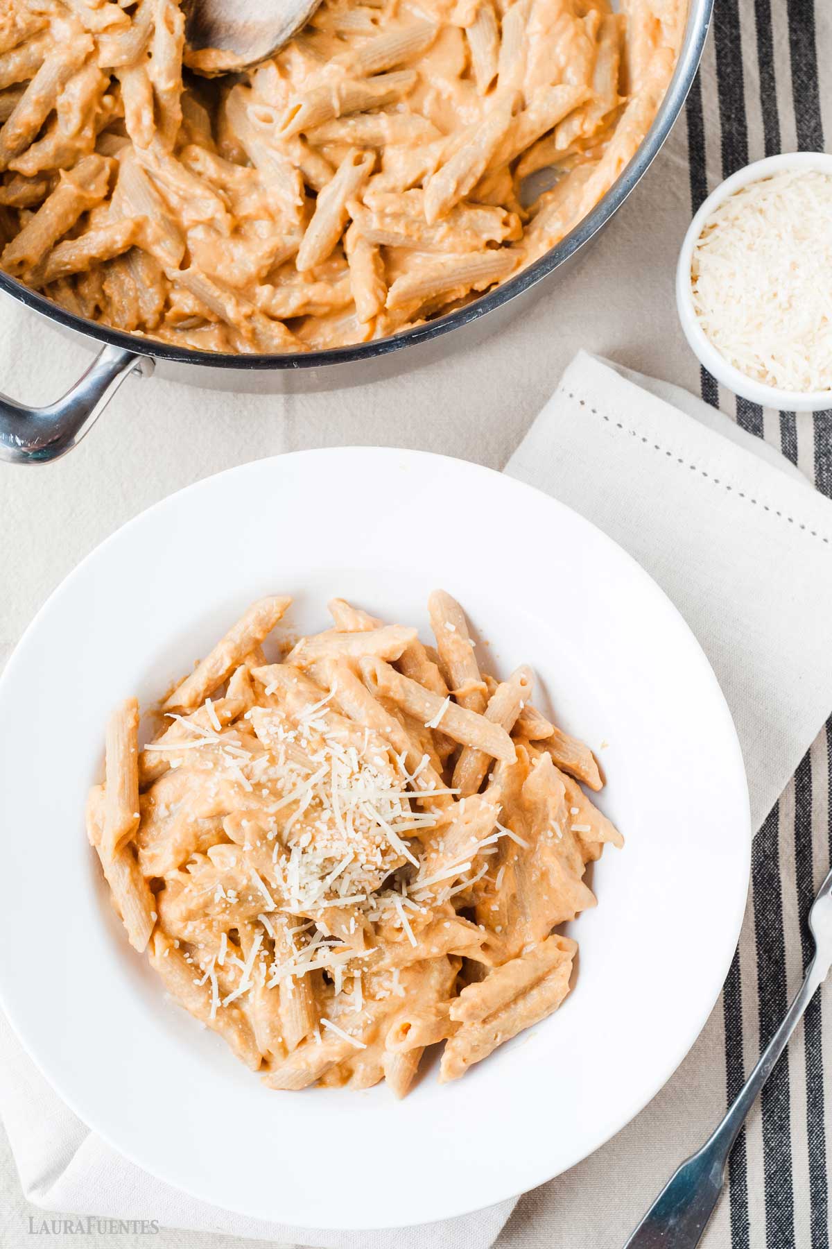 una sartén y un plato, ambos con pasta de calabaza cremosa, junto a un bowl pequeño con queso rallado.