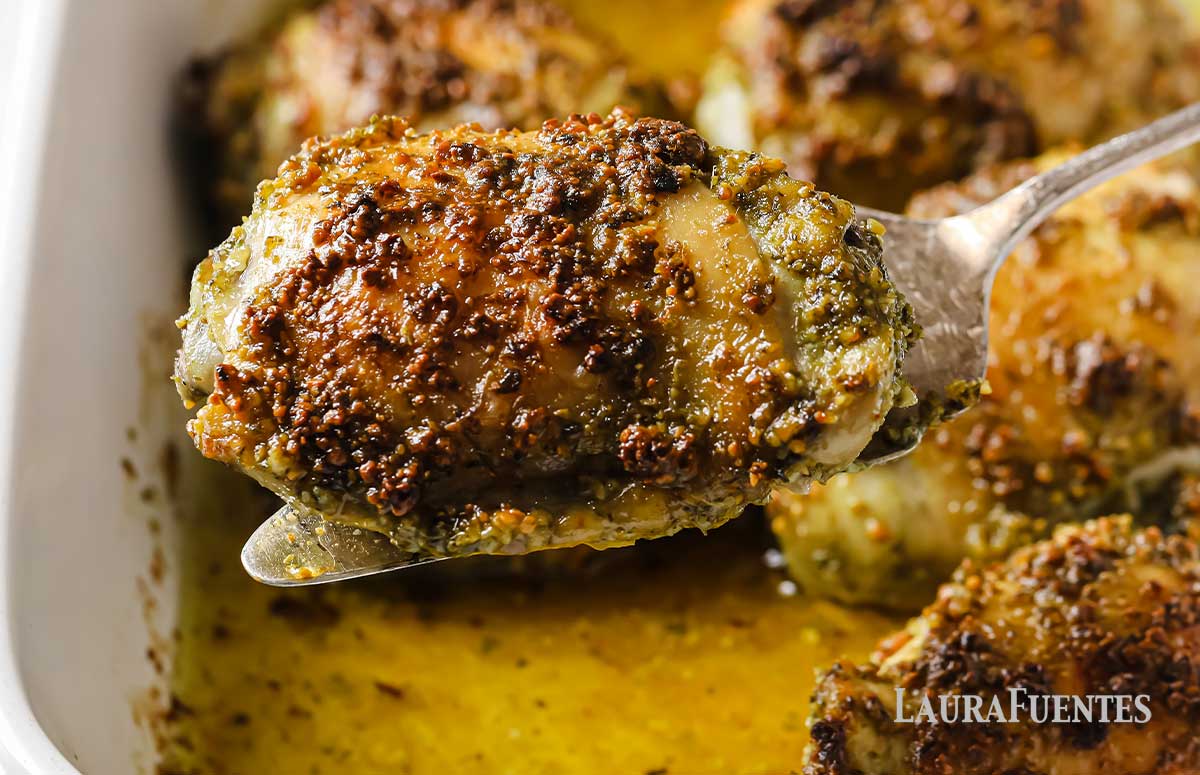 una cuchara sosteniendo un muslo de pollo con pesto, detrás una fuente con más muslos de pollo
